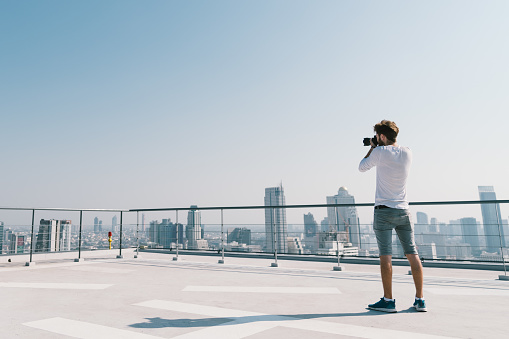 Young white Caucasian man taking cityscape photo on building rooftop on sunny day. Photography hobby, gadget technology, or leisure activity concept. With copy space on blue sky