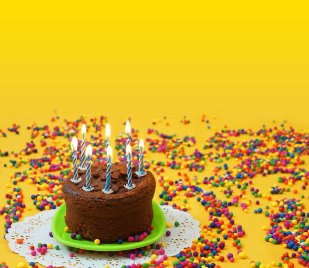 istock Chocolate birthday cake with lighted blue and white candles, on a small green plate surrounded by candy balls strewn on a yellow background. Image with copy space 911548428