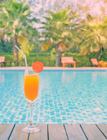 Orange juice with carrot slice in cocktail glass at outdoor swimming pool, summer tropical holiday concept. Vintage filtered image.