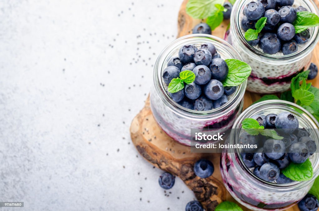 Chia pudding with blueberry, mint leaves  and jam Chia pudding with blueberry and jam in glass jars on a wooden board and light stone background. Horizontal image, copy space, high angle view Blueberry Stock Photo