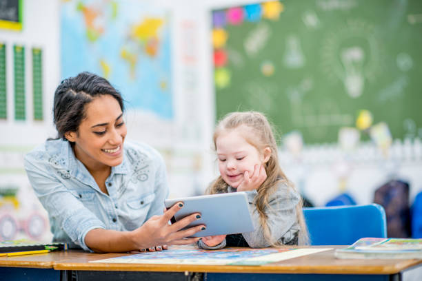 Watching A Video Together A girl with down syndrome is in a classroom with her teacher. The teacher is holding a tablet computer to let the girl watch a video. They are both smiling. special education stock pictures, royalty-free photos & images