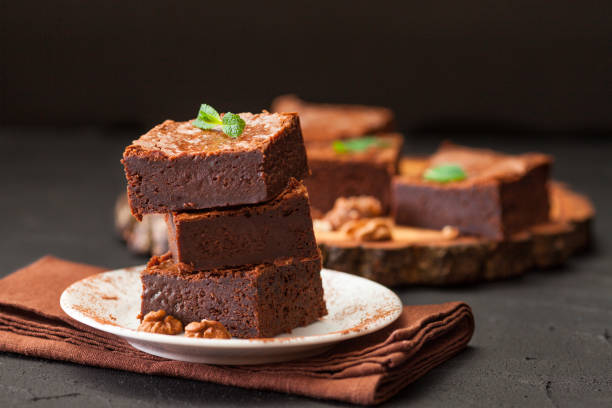 schokoladen-brownie quadratische stücke im stapel auf weißen teller mit walnüssen, dekoriert mit minze und kakao auf schwarzem hintergrund. köstliches dessert. düstere stimmung. fotografie hautnah. selektiven fokus - close up cookie gourmet food stock-fotos und bilder