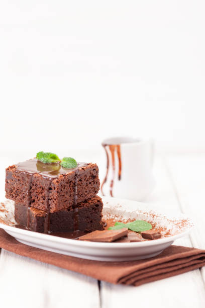 istock Chocolate brownie square pieces in stack on white plate decorated with mint leaves and cocoa powder on white vintage wooden background. American traditional delicious dessert. Close up photography 911640888