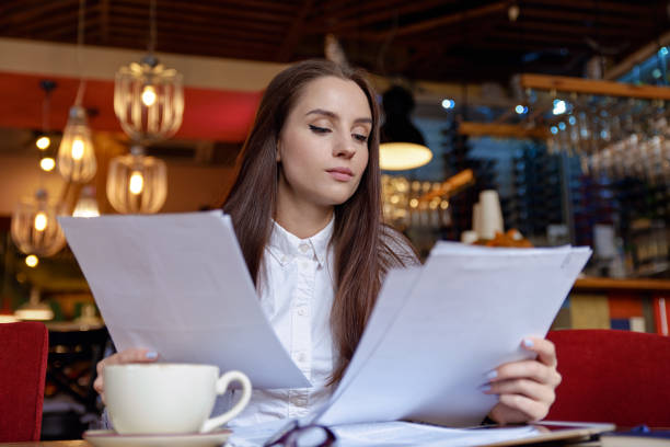 dame élégante affaires confiante attractif avec de longs cheveux étudie les documents qui les détient avec les deux mains devant elle, autour d’une tasse de café, assis au café-restaurant - paper document pen long hair photos et images de collection