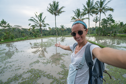 Cheerful girl standing by a rice terrace in Bali takes a selfie portrait. People travel joy concept