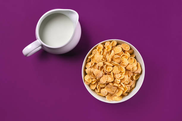 Bowl with corn flakes, jug of milk on purple background, top view stock photo