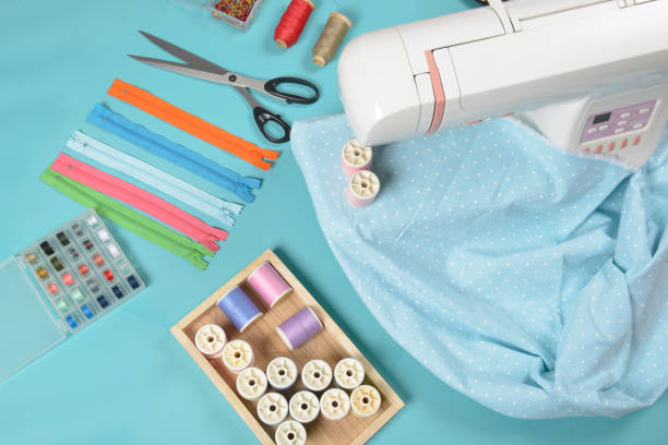 istock Fabrics on sewing machine amid the scissors, shirt buttons, zipper, pin and colorful thread rolls for sewing on blue background, Sewing and needlework concept. 911687038
