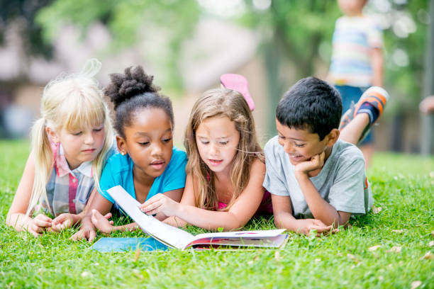 Kids Reading Three young girls and a boy are outdoors during summer. They are laying in the grass and reading a book together. boy and girl reading stock pictures, royalty-free photos & images