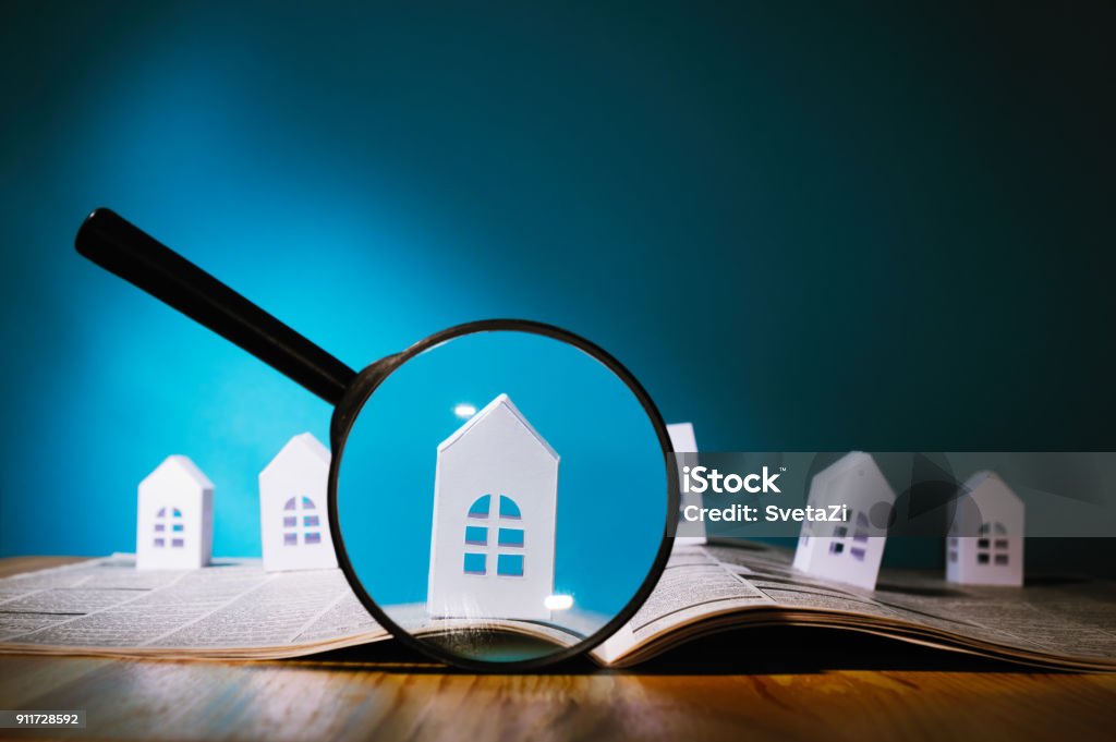 Paper house under a magnifying lens Magnifying glass in front of an open newspaper with paper houses. Concept of rent, search, purchase real estate. House Stock Photo