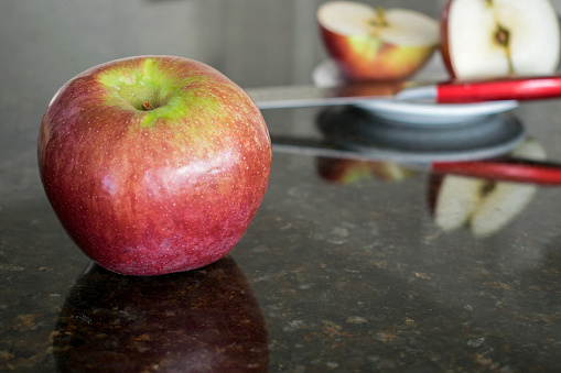 Apple in front with knife and cut apple on plate behind with reflections on granite counter.