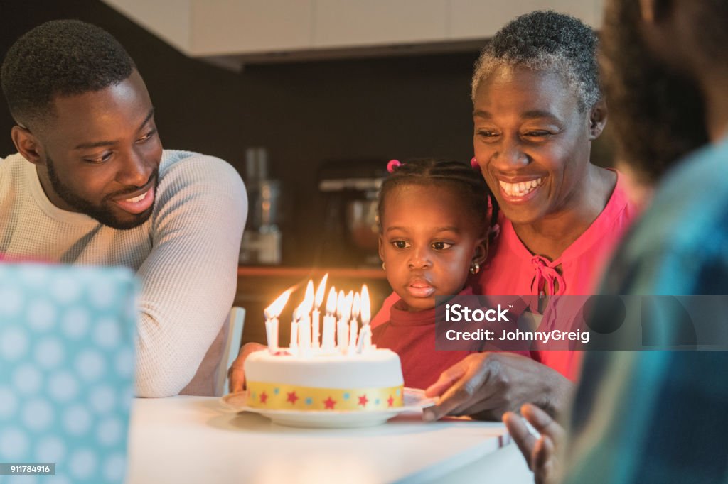 Grandmother with birthday cake and granddaughter sitting on lap staring Three generation African family celebrating birthday, young girl looking at cake on her grandmother's lap, father looking towards daughter Birthday Stock Photo