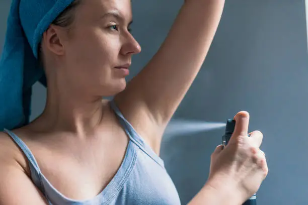 Beautiful woman in bath towel is applying deodorant while standing in bathroom on underarm after having a shower. Photo of female applying water spray while standing at home during the day.