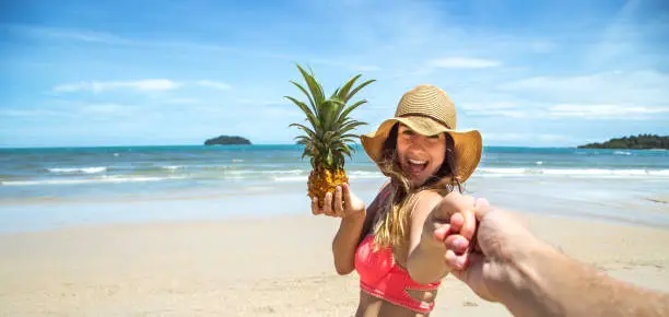 Photo of beautiful girl in swimsuit and pineapple walks on the beach holding the hand of the guy