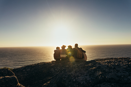 Rear view of friends sitting together on mountain top and enjoying the sunset over sea. Men and women relaxing on mountain top during sunset.