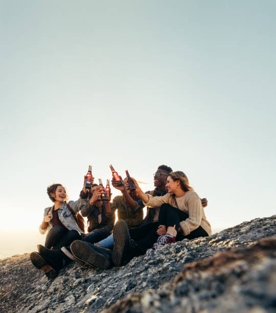 Group of friends on a mountain partying with beers. Young men and women toasting beers during sunset.
