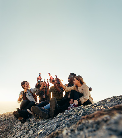 Group of friends on a mountain partying with beers. Young men and women toasting beers during sunset.