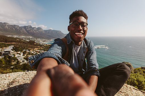 Smiling african guy sitting on top of a mountain taking selfie against seascape. Man enjoying his holiday and making self portrait against beautiful landscape.