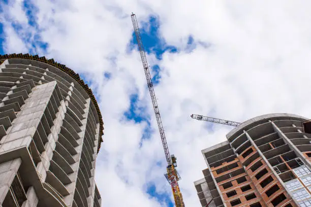 A construction crane on the background of the construction of two houses or skyscrapers and blue sky with clouds bottom view