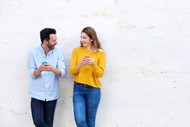 riant debout couple par du mur blanc comprenant des téléphones mobiles - côte à côte photos et images de collection