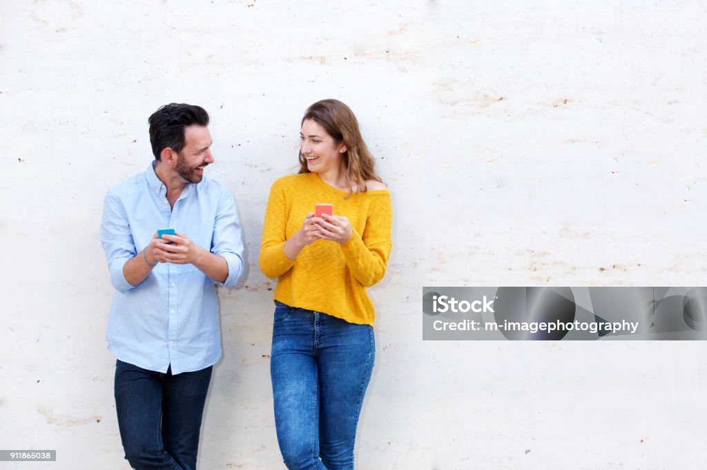 Laughing couple standing by white wall holding mobile phones Portrait of laughing couple standing by white wall holding mobile phones Friendship Stock Photo