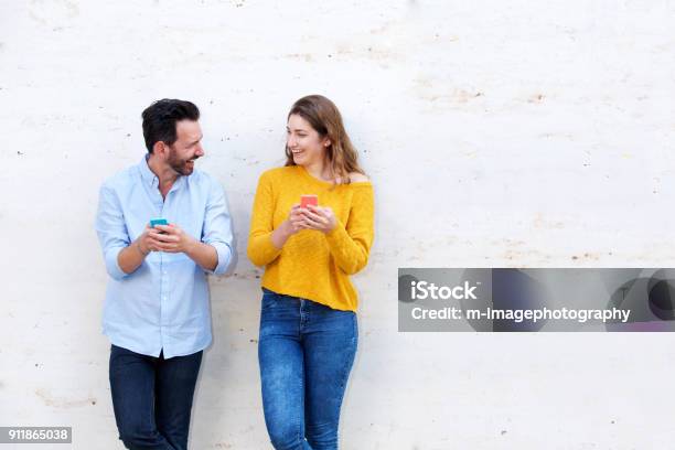 Laughing Couple Standing By White Wall Holding Mobile Phones Stock Photo - Download Image Now