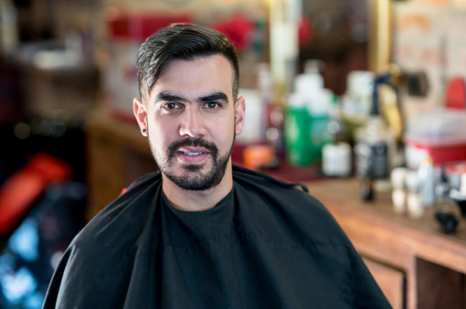 Portrait of a happy Latin American man getting a haircut at a hair salon and wearing a cape while looking at the camera