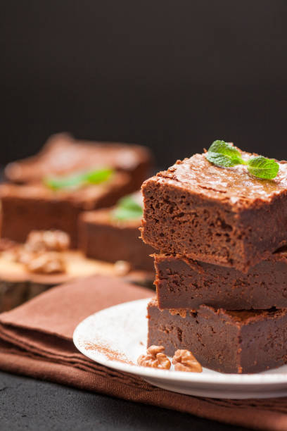 istock Chocolate brownie square pieces in stack on white plate with walnuts, decorated with mint leaves and cocoa on black background. Delicious dessert. Dark mood. Close up photography. Selective focus 911892190