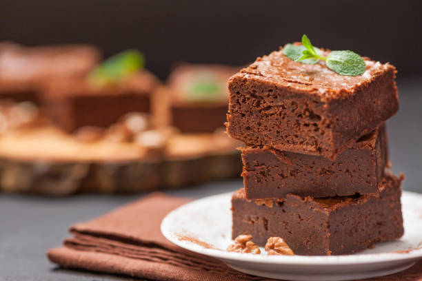 istock Chocolate brownie square pieces in stack on white plate with walnuts, decorated with mint leaves and cocoa on black background. Delicious dessert. Dark mood. Close up photography. Selective focus 911892204