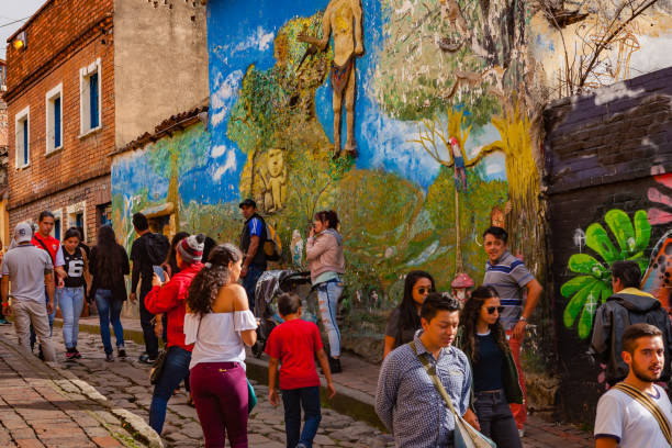 Bogotá, Colombia - Tourists And Local Colombians On The Narrow, Cobblestoned Calle del Embudo In The Historic La Candelaria District Of The Andes Capital City Bogotá, Colombia - May 28, 2017: A few tourists and some local Colombian families seen on the broader end of the, colourful, cobblestoned Calle del Embudo which is at the opposite end of the Plaza del Chorro de Quevedo. The Street gets its name from its funnel shape: Embudo translates to Funnel in English. It is shaped like a funnel. The street was constructed over 450 years ago when people usually travelled on horseback; it is well known for its Street Art, many of which feature the legends of the Pre-Colombian era. Photo shot in the late afternoon sunlight; horizontal format. Camera: Canon EOS 5D MII. Lens: Canon EF 24-70 F2.8L USM. calle del embudo stock pictures, royalty-free photos & images