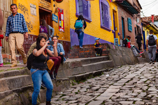Bogotá, Colombia - Tourists And Local Colombians On The Narrow, Cobblestoned Calle del Embudo In The Historic La Candelaria District Of The Andes Capital City Bogotá, Colombia - May 28, 2017: A few tourists and some local Colombians seen on the broader end of the, colourful, cobblestoned Calle del Embudo which is at the opposite end of the Plaza del Chorro de Quevedo. The Street gets its name from its funnel shape: Embudo translates to Funnel in English. It is shaped like a funnel. The street was constructed over 450 years ago when people usually travelled on horseback; it is well known for its Street Art, many of which feature the legends of the Pre-Colombian era. Photo shot in the late afternoon sunlight; horizontal format. Camera: Canon EOS 5D MII. Lens: Canon EF 24-70 F2.8L USM. calle del embudo stock pictures, royalty-free photos & images
