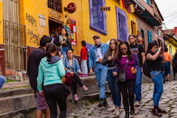 Bogotá, Colombia - Tourists And Local Colombians On The Narrow, Cobblestoned Calle Del Embudo In The Historic La Candelaria District Of the Andes Capital City Bogotá, Colombia - May 28, 2017: A few tourists and some local Colombian families seen on the broader end of the, colourful, cobblestoned Calle del Embudo which is at the opposite end of the Plaza del Chorro de Quevedo. The Street gets its name from its funnel shape: Embudo translates to Funnel in English. It is shaped like a funnel. The street was constructed over 450 years ago when people usually travelled on horseback; it is well known for its Street Art, many of which feature the legends of the Pre-Colombian era. Photo shot in the late afternoon sunlight; horizontal format. Camera: Canon EOS 5D MII. Lens: Canon EF 24-70 F2.8L USM. calle del embudo stock pictures, royalty-free photos & images