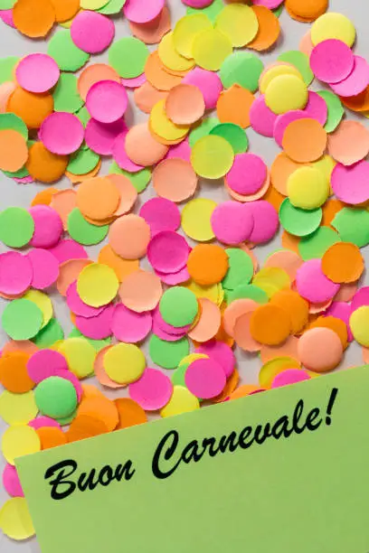 Carnival party background concept, space for text. Written the words in italian language: Buon  Carnevale. Colorful confetti spread over table. Warm colors: pink, yellow and orange. Overhead, flat lay, vertical orientation.