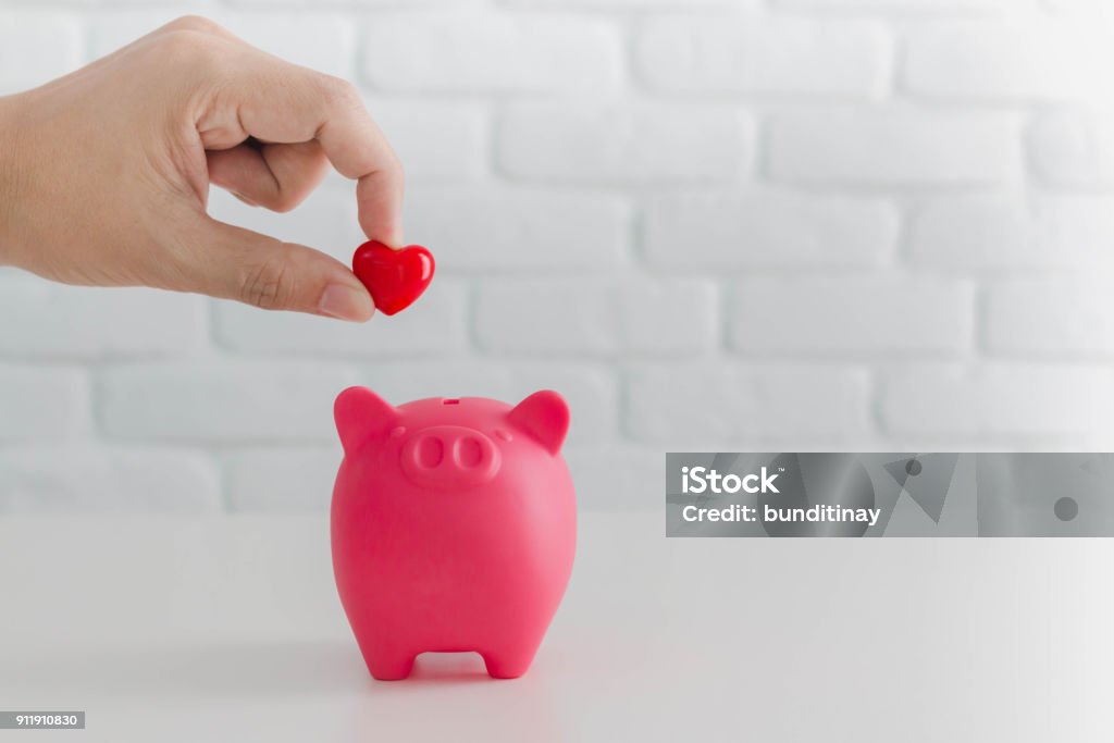 Man's hand putting red heart in to piggy bank metaphor saving love for lover or family in every day.Concept of happy relationship. Currency Stock Photo