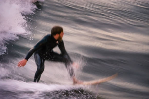 The lone surfer is catching a wave on his surfboard, and is wearing a wetsuit to protect him from the frigid waters of the Pacific Ocean. Seal Beach in Orange County, is a popular destination for Southern Californian surfers. Photo was taken off the Seal Beach pier. A slow shutter speed along with panning was used to create the motion blur, and render the surfer unrecognizable.