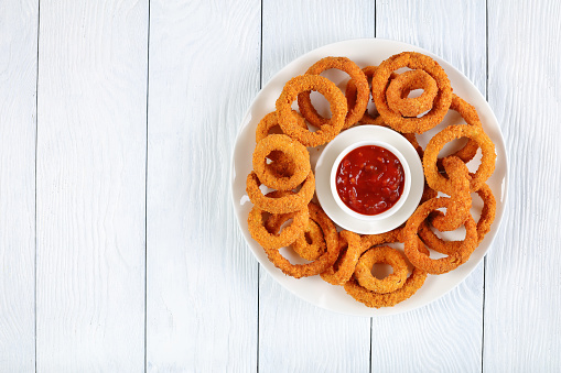 delicious golden battered, breaded and deep fried crispy onion rings served on white platter with ketchup in center, view from above