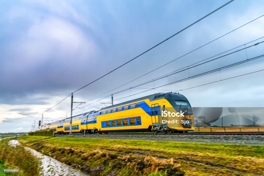 Train driving in a rural landscape during a dark day Yellow and blue Dutch passenger train driving through a rural landscape on a dark and rainy fall day. Image with speed effect from the motion blur. Train - Vehicle Stock Photo