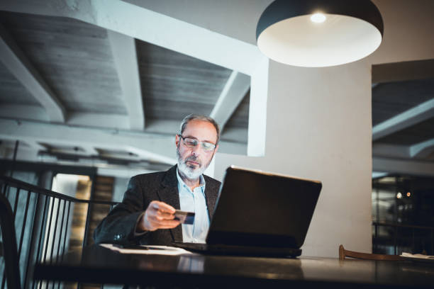 istock Senior businessman with beard making credit card purchase using laptop in high end restaurant 911978164