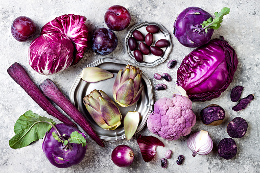Raw purple vegetables over gray concrete background. Cabbage, radicchio salad, olives, kohlrabi, carrot, cauliflower, onions, artichoke, beans, potato, plums. Top view, flat lay.