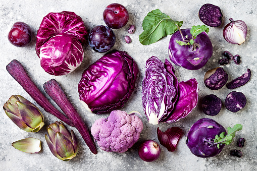 Raw purple vegetables over gray concrete background. Cabbage, radicchio salad, kohlrabi, carrot, cauliflower, onions, artichoke, beans, potato, plums. Top view, flat lay.