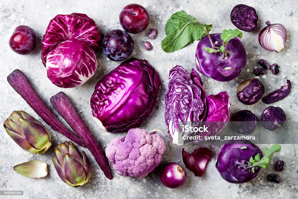 Raw purple vegetables over gray concrete background. Cabbage, radicchio salad, olives, kohlrabi, carrot, cauliflower, onions, artichoke, beans, potato, plums. Top view, flat lay. Raw purple vegetables over gray concrete background. Cabbage, radicchio salad, kohlrabi, carrot, cauliflower, onions, artichoke, beans, potato, plums. Top view, flat lay. Purple Stock Photo