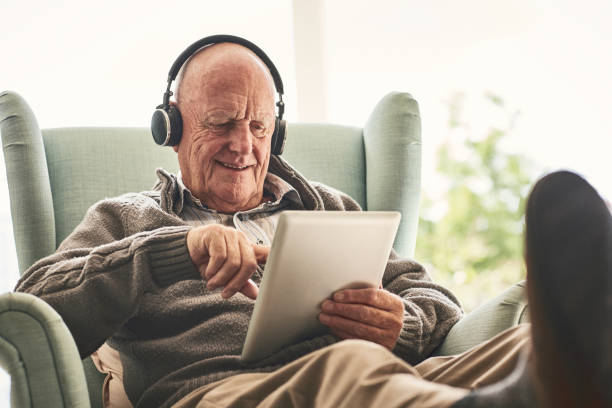 Happy elderly man at home using digital tablet Happy elderly man relaxing on a chair at home and listening to music on digital tablet with headphones armchair photos stock pictures, royalty-free photos & images