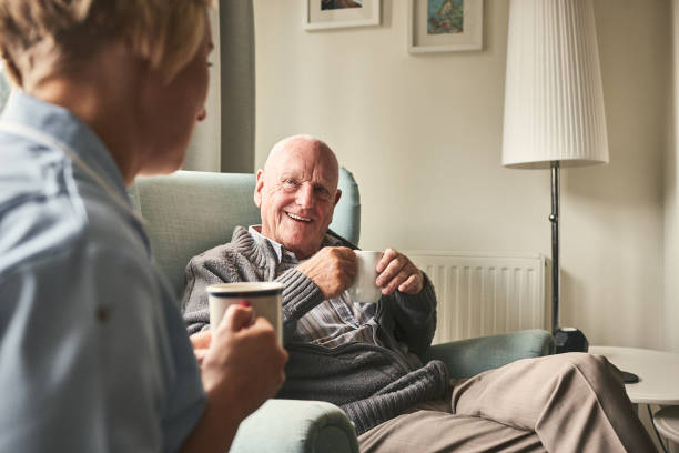 Smiling senior man talking to female caregiver Smiling senior man with cup of coffee talking to female caregiver sitting in front at care home seniors talking stock pictures, royalty-free photos & images