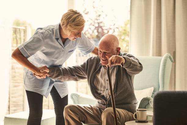 Female healthcare worker supporting senior man at care home Smiling senior man with female healthcare worker. Home carer supporting old man to stand up from the chair at care home. walking stick stock pictures, royalty-free photos & images