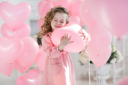 A little girl of 6 years with long curly hair, dressed in a pink dress and white tights, a beautiful smile, sits alone in a large bright room with lots of pink balloons in the shape of a heart. Valentine's Day and party celebration.