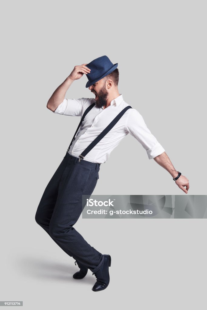 Perfect in every way. Full length of handsome young man in suspenders adjusting his hat and making a face while dancing against grey background Dancing Stock Photo