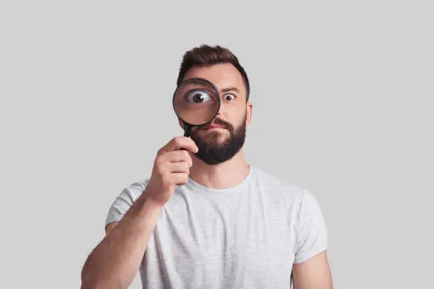 Playful young man applying magnifying glass while standing against grey background