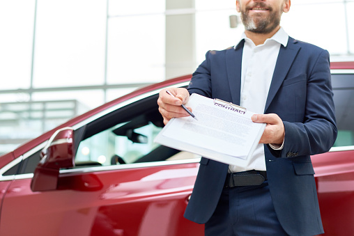 Mid section portrait of smiling bearded salesman handing purchase contract to client buying brand new car in luxury showroom, copy space
