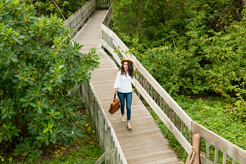 This is a horizontal, color photograph of a beautiful, Millennial woman of Israeli descent walking with her acoustic guitar on a wooden board walk through a lush, green park in Miami, Florida, USA. The young American woman is photographed from a high angle view. Mangrove trees line the footpath.