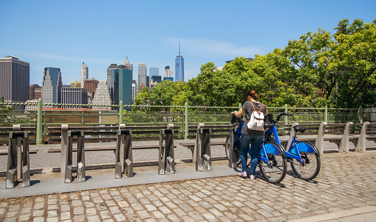 Back view of unrecognizable woman with backpack taking a bike in front of Manhattan skyline, in New York City