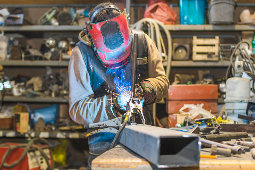 young man at the Metal shop with the mask on his face welding a bar in progress surrounded by tools and metal